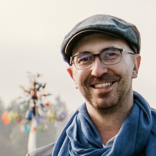 Kaelig Deloumeau-Prigent smiling outdoors, wearing a flat cap, glasses, and a blue scarf.